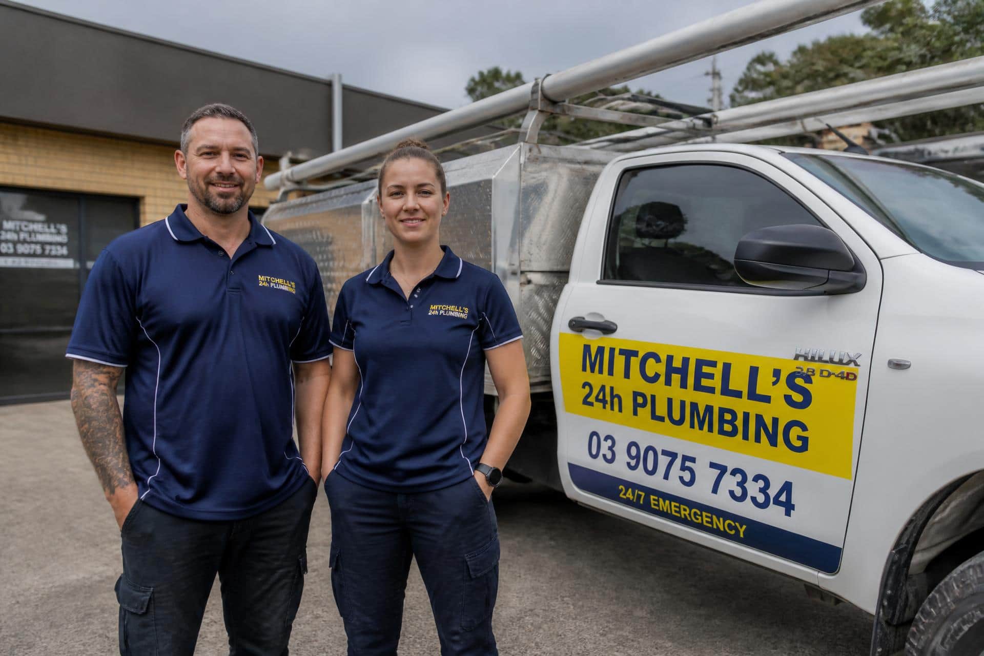 Two Mitchell's 24H Plumbing plumbers standing in front of their service ute, signwritten with 03 9075 7334